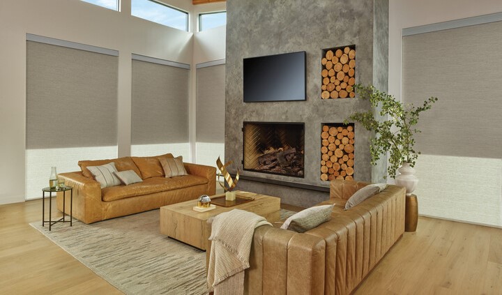 A living room with a tan leather sofa, a stone fireplace with a television above it, and large windows with motorized blinds..